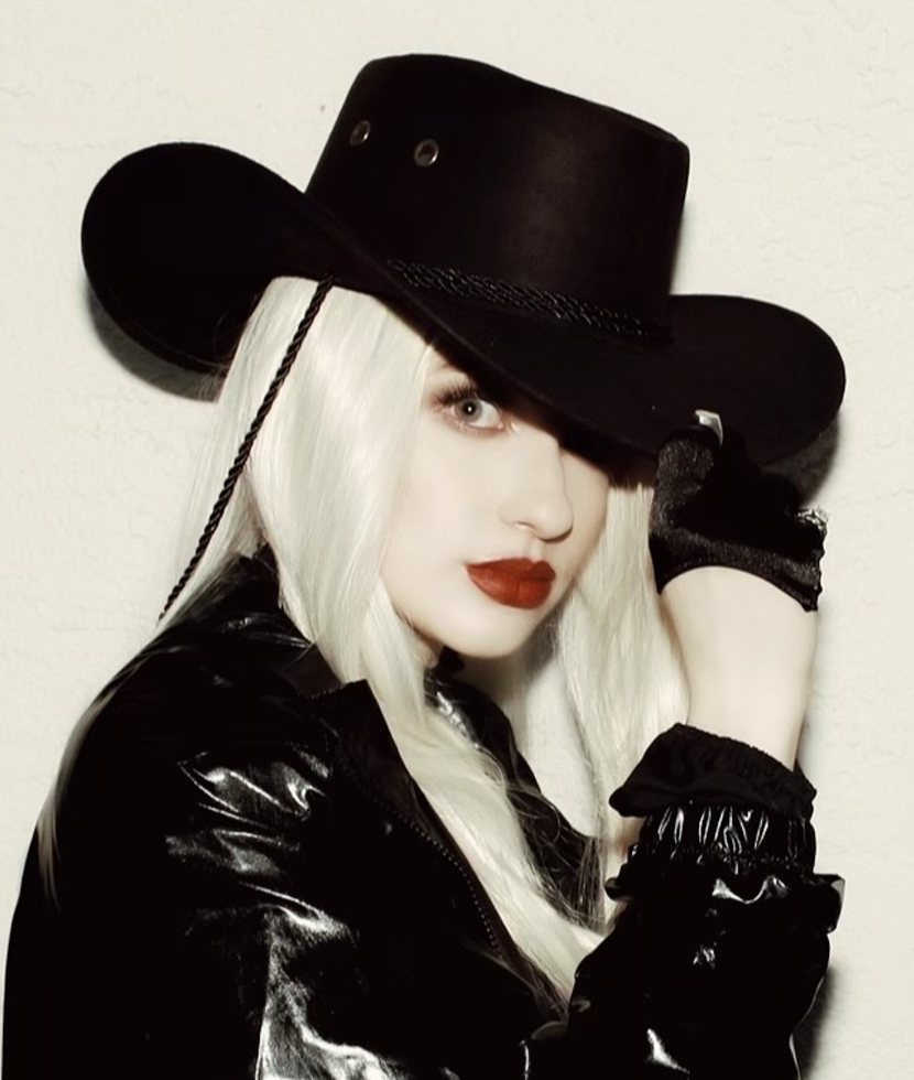 Artist in a black cowboy hat, white-blonde hair and a patent leather jacket, photographed against a pale wall.