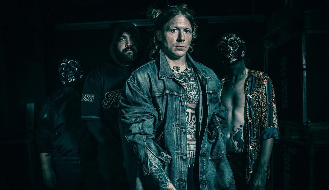 Four-piece band in the dark backline of a venue: the frontman in a denim jacket up front, flanked by two masked members and a hooded bandmate.