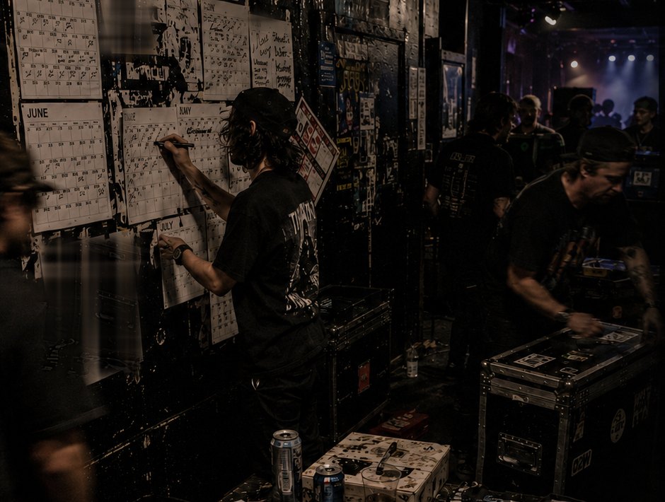 Artist and touring crew updating printed release calendars backstage while equipment moves through a small venue.